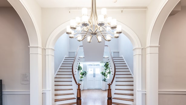 A grand staircase with white railings and a chandelier, leading to a door with potted plants. - The Blackburn Inn and Conference Center (Staunton)