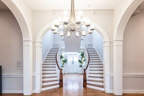 A grand staircase with white railings and a chandelier, leading to a door with potted plants. - The Blackburn Inn and Conference Center (Staunton)