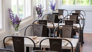 A dining area with tables and chairs, vases with flowers, and large windows. - The Blackburn Inn and Conference Center (Staunton)