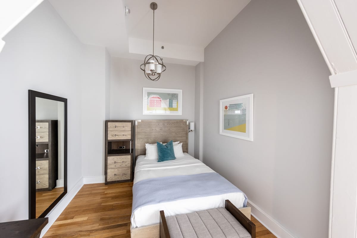 Une chambre avec un plancher en bois, un lit recouvert d’une couvre-lit à rayures blanches et bleues, une tête de lit en bois, une table de chevet et un miroir sur le mur de gauche.