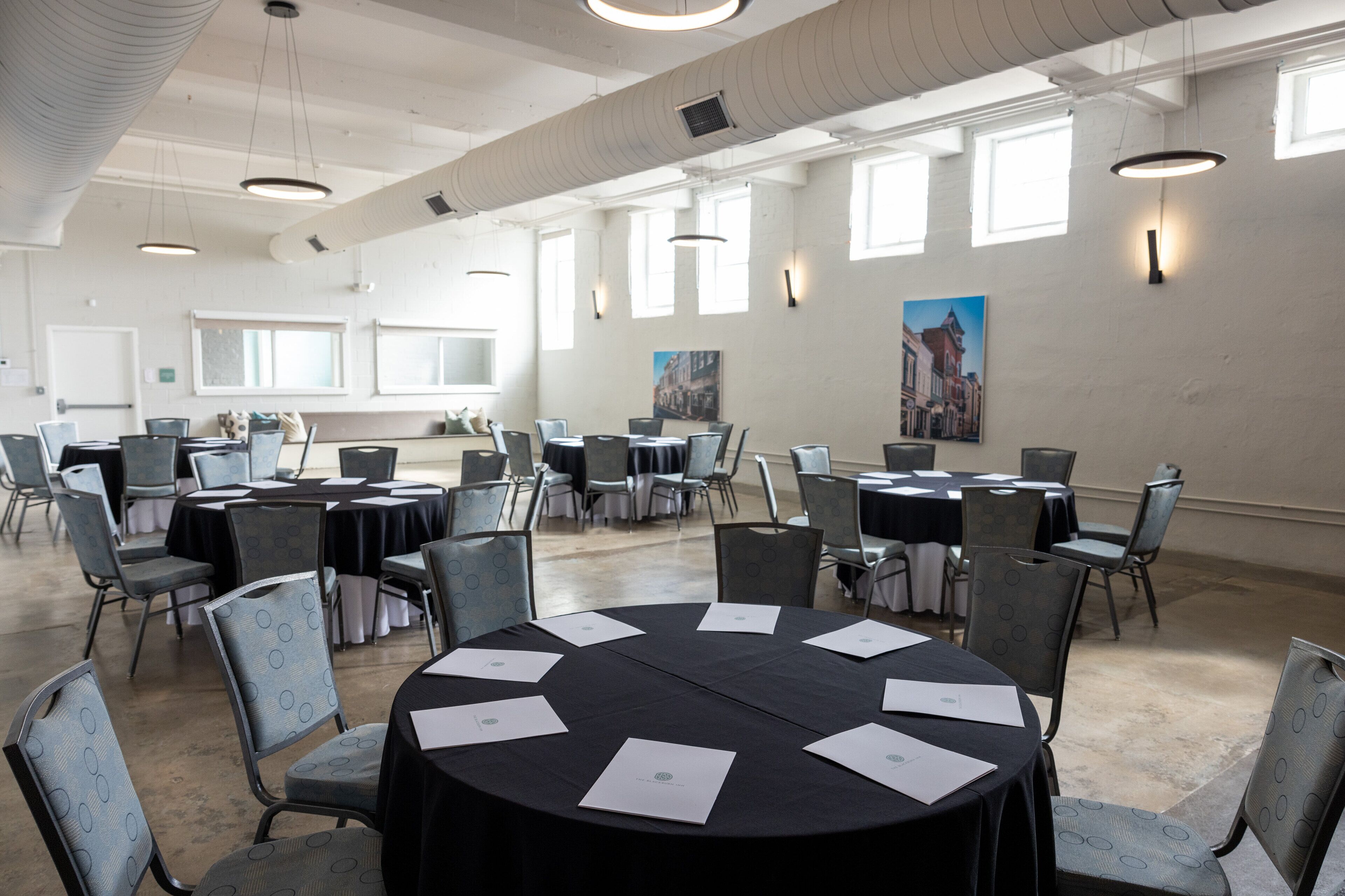A conference room with round tables, chairs, and papers, featuring large windows and artwork on the walls.