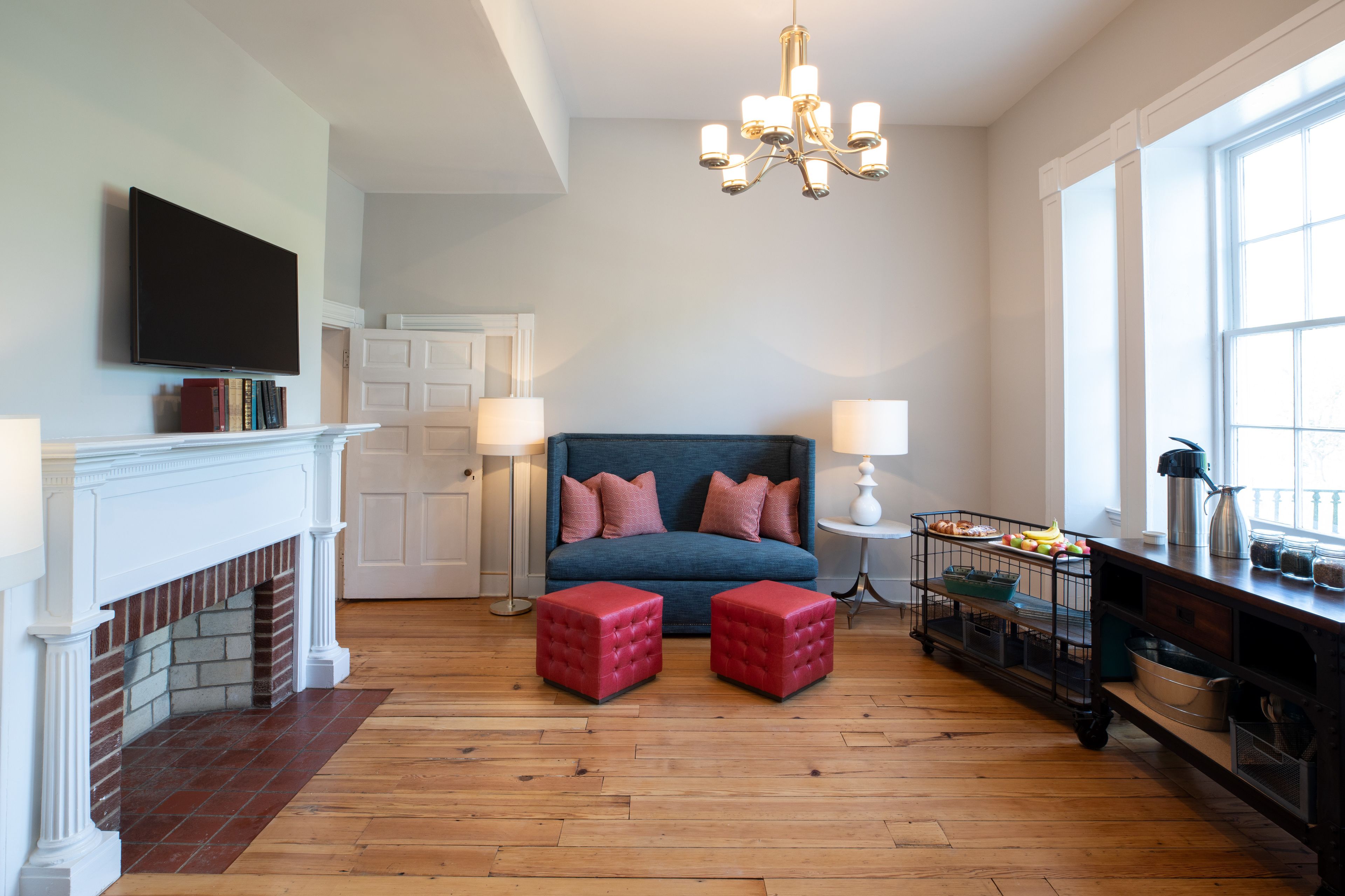 A bedroom with a fireplace, a bed with a blue headboard, two red ottomans, a chandelier, and a window with a view.