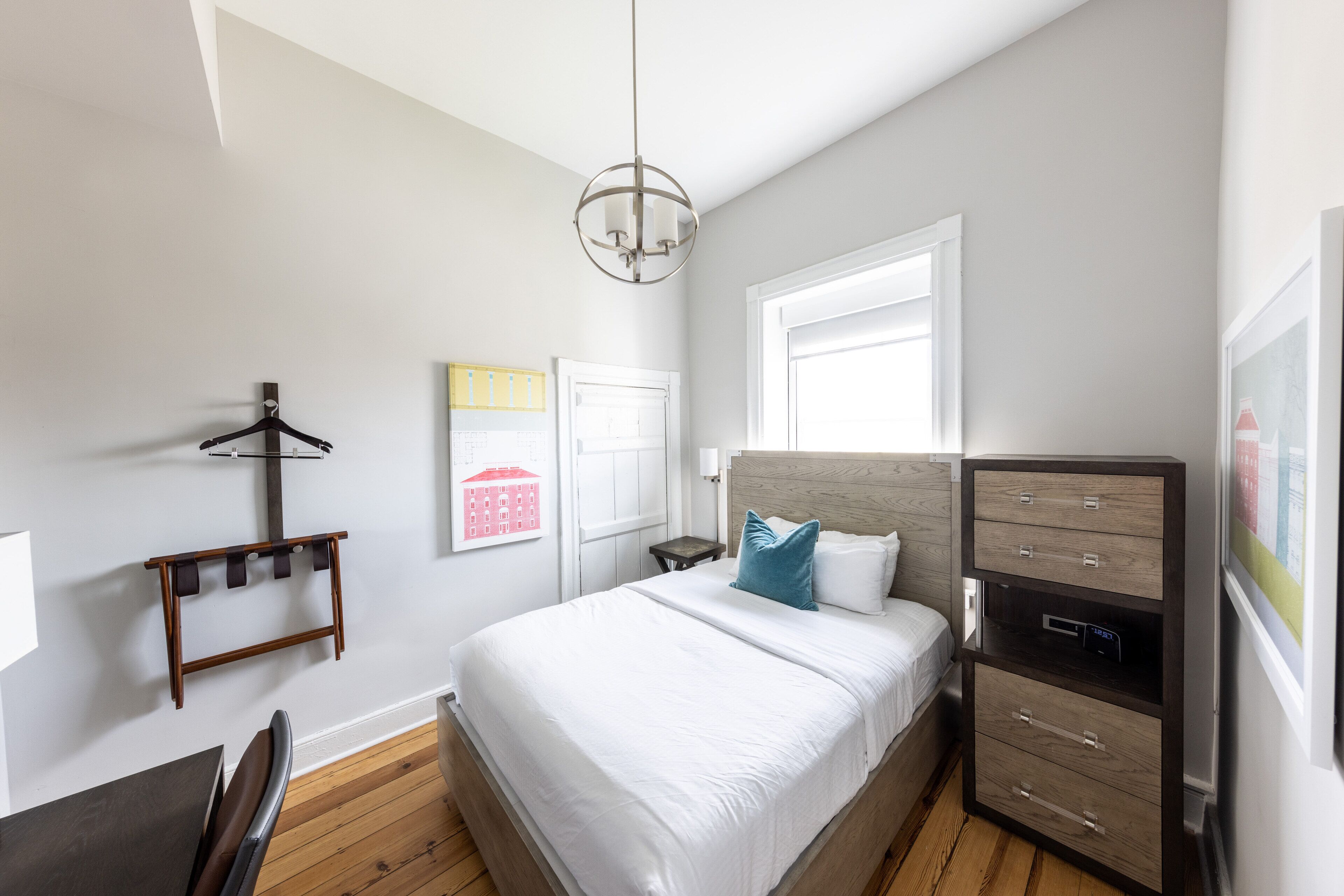A bedroom with a bed, a dresser, a chair, a coat rack, and a wall-mounted artwork.