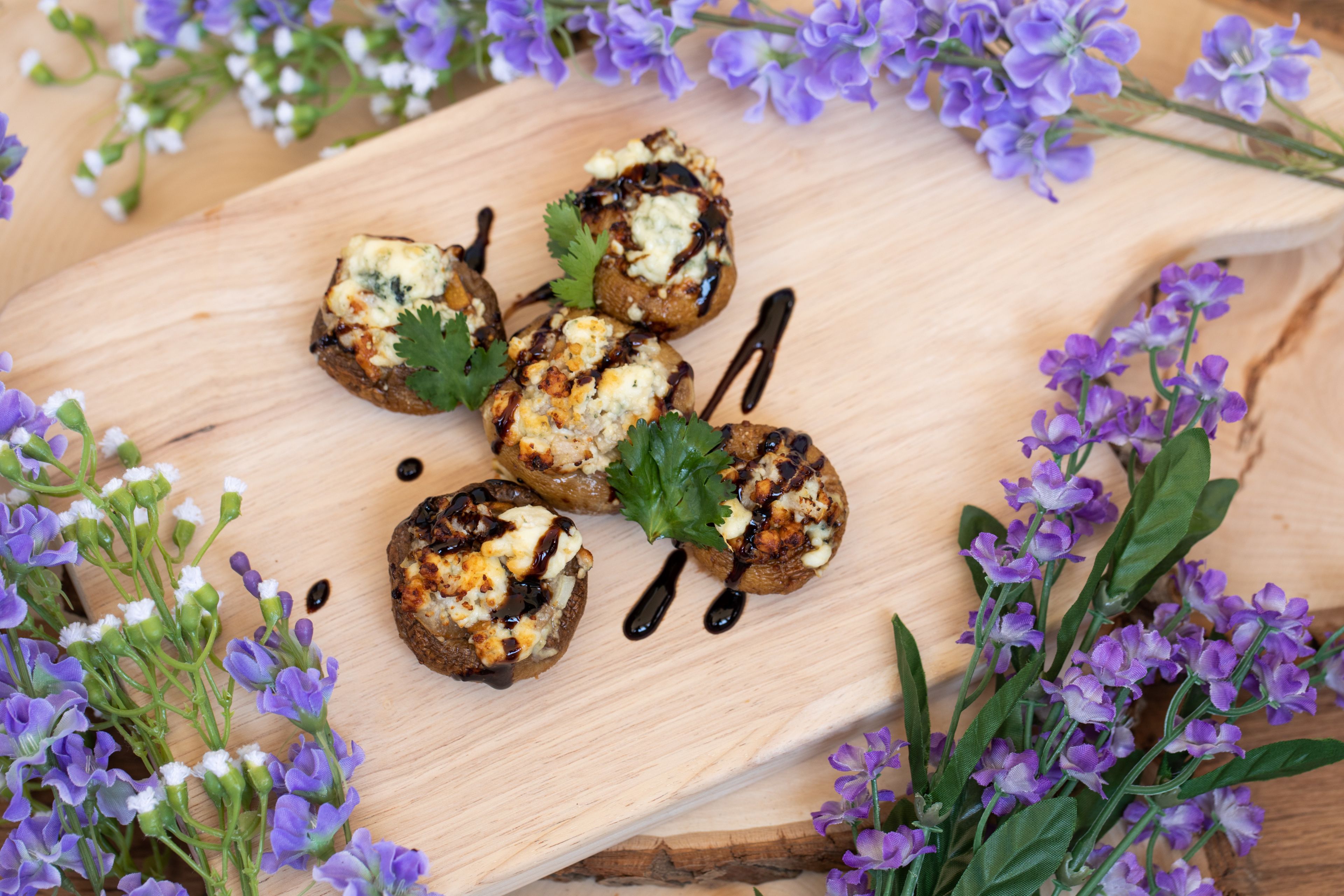A wooden board with grilled mushrooms garnished with cheese and herbs, surrounded by purple flowers.