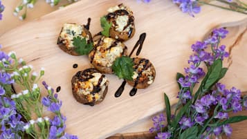 Una tabla de madera con champiñones a la parrilla, adornados con queso y hierbas, rodeada de flores moradas.