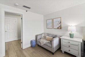 A room with a grey sofa, a white bedside table with a lamp, and a wall with two framed pictures.