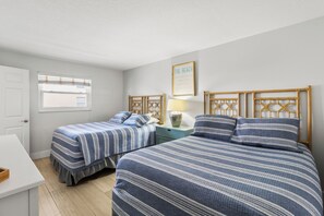 A bedroom with two single beds, a bedside table with a lamp, a framed quote on the wall, and a window with blinds.