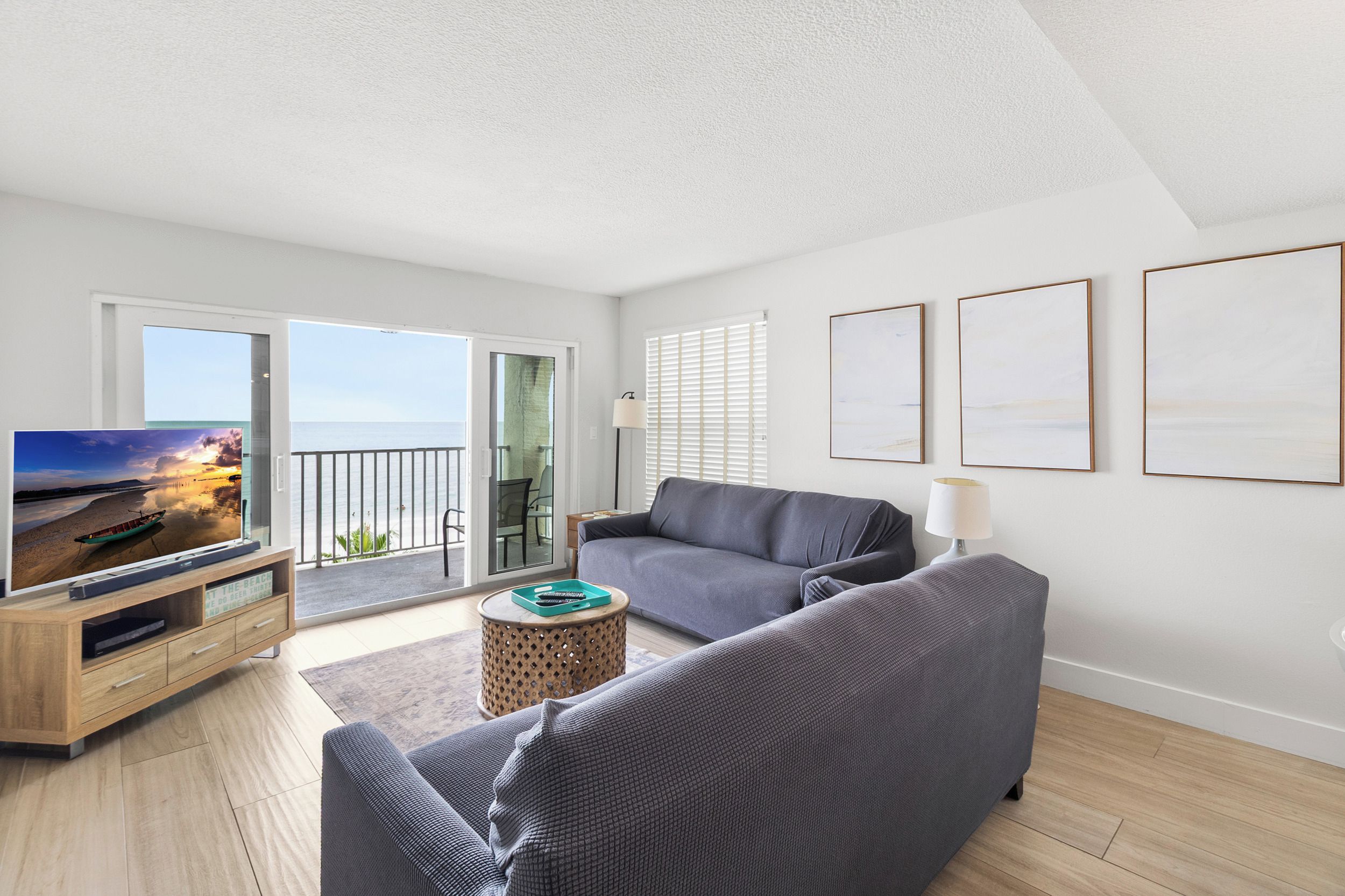Un salón moderno con un sofá gris, un televisor sobre un mueble de madera y un balcón con vistas al mar.