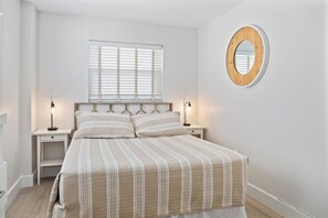 A bedroom with a bed, two bedside tables, a window with blinds, and a round mirror on the wall.