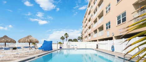 Une piscine entourée d’une clôture et d’un bâtiment doté de balcons, avec des chaises longues et des parasols à proximité.