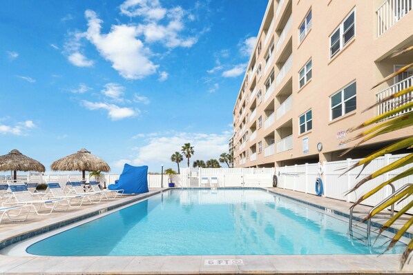 A swimming pool with lounge chairs and umbrellas, surrounded by a fence and a building with balconies.