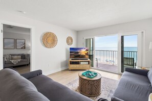 A modern living room with a large TV, grey sofas, a wooden coffee table, and a balcony with a view of the sea.