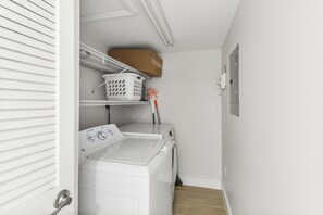 A laundry room with a washing machine, a shelf with a basket, and a wall-mounted electrical outlet.