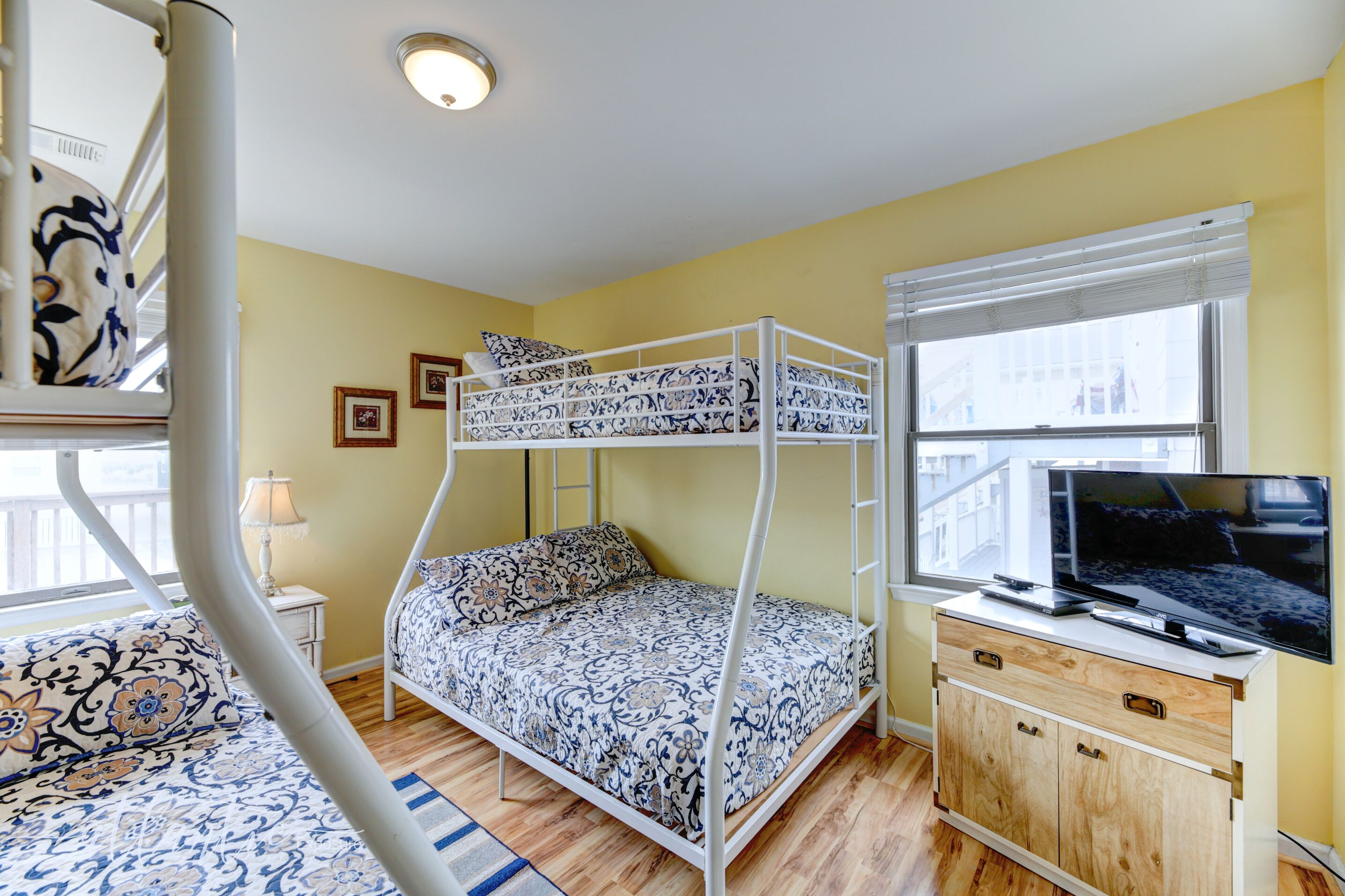 Una cama tipo litera con colcha estampada, un televisor sobre un mueble de madera y una ventana con persianas.