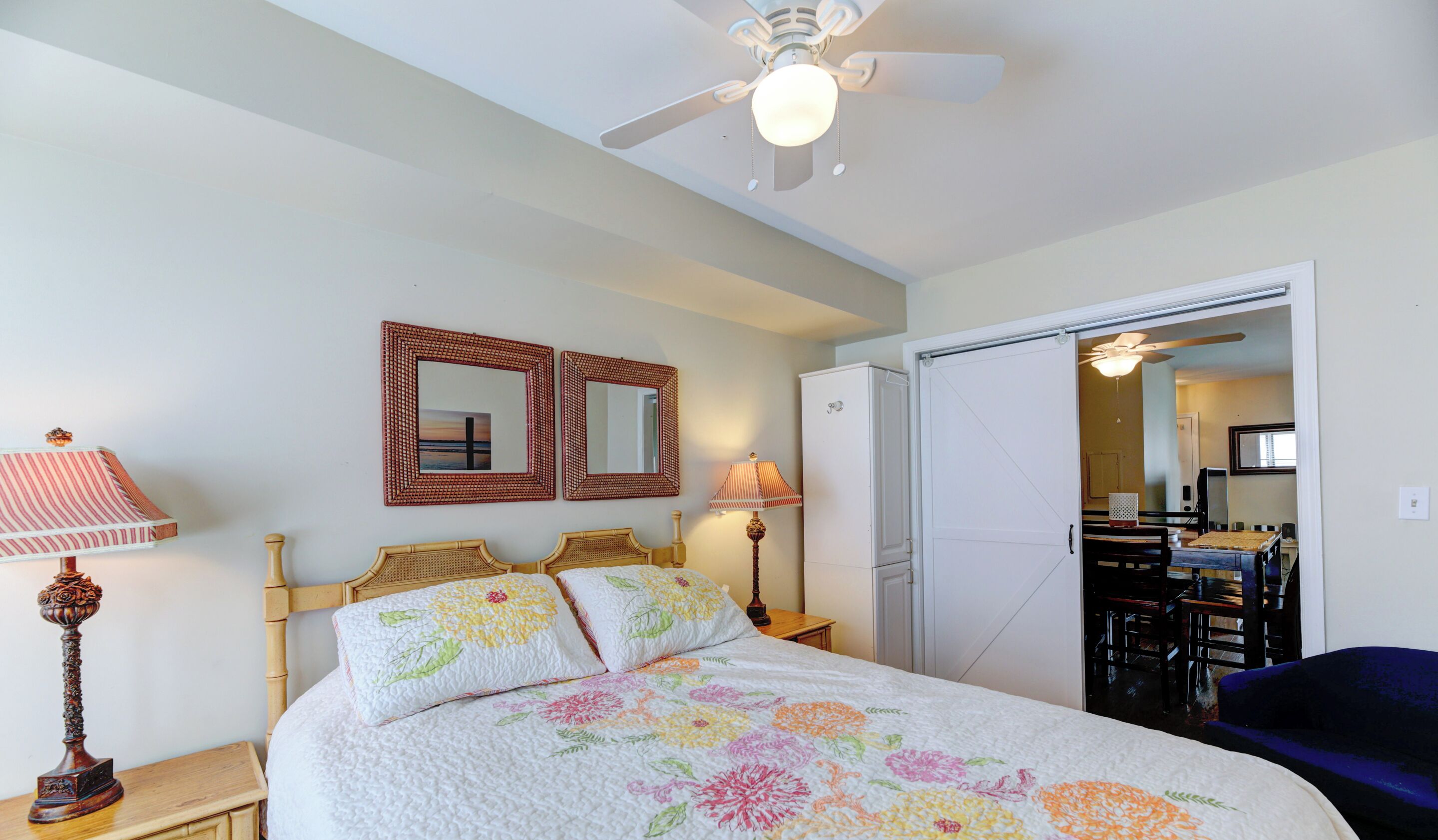 Un dormitorio con una cama, dos espejos en la pared, un ventilador de techo y una puerta que da a un comedor.