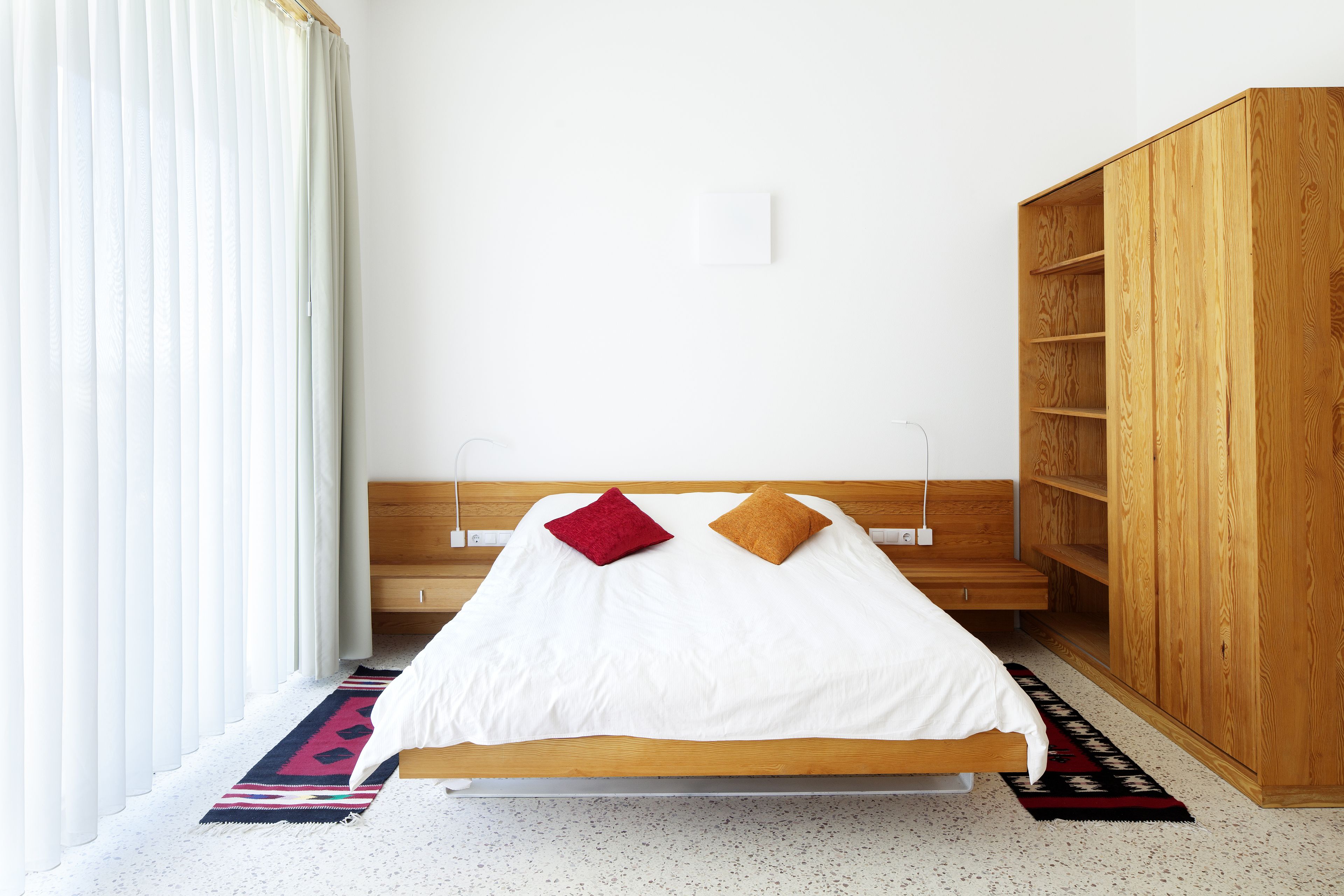 Ein ordentlich bezogenes Bett mit Holz-Kopfteil, ein großer Kleiderschrank und ein gemustertes Teppich auf einem gefleckten Boden.