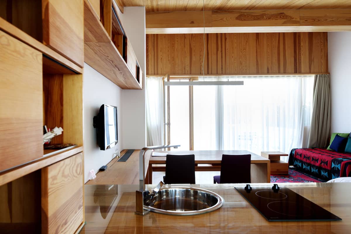A modern kitchen with wooden cabinets, a dining area with chairs, a large window, and a bed with a colorful blanket.