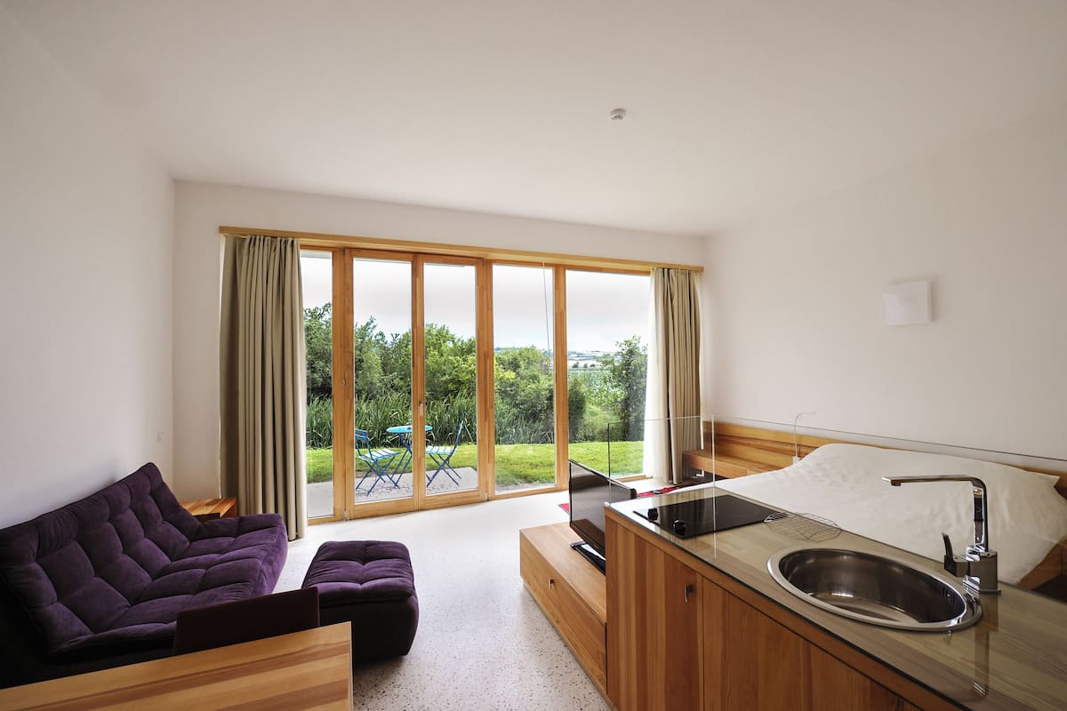 A modern hotel room with a sofa, a small table, a bed with a built-in sink, and large sliding glass doors to a garden.