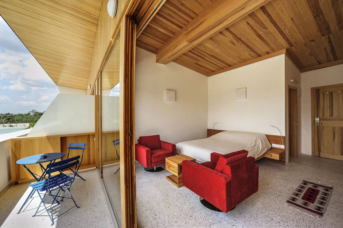 A modern bedroom with a large bed, red chairs, a small table, and a balcony with a view.