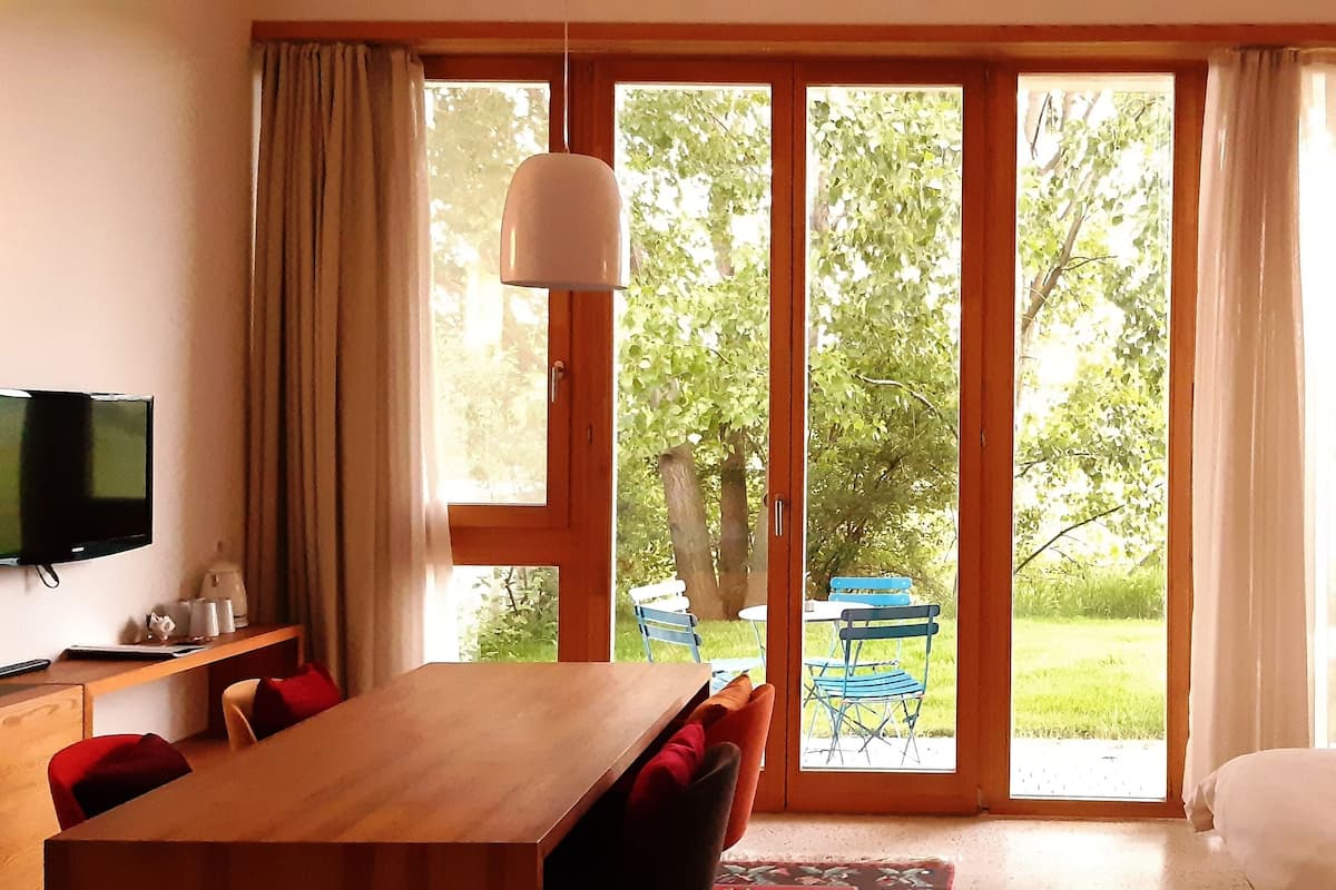 A room with a wooden table, chairs, a bed, a TV, and a sliding glass door leading to a garden.