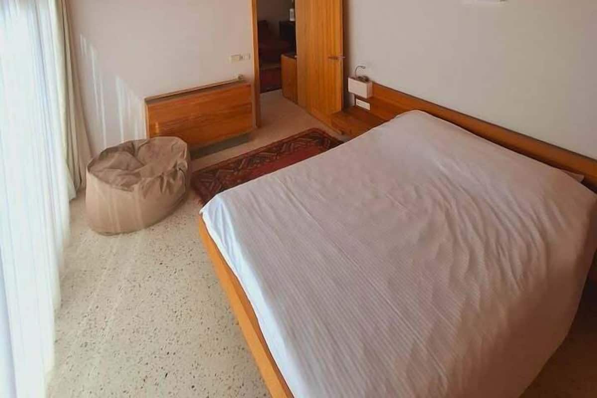 A bedroom with a wooden ceiling, a bed, a bedside table, a nightstand, a wardrobe, and a window with curtains.