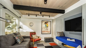 Una sala de estar moderna con sofá, sillón, reposapiés y un sofá cama azul. Hay un televisor grande montado en la pared, un reloj y un cartel.