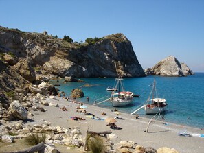 Una platja rocosa amb vaixells amarrats a prop de la riba, un mar blau i translúcid, i un paisatge de muntanyes al fons.