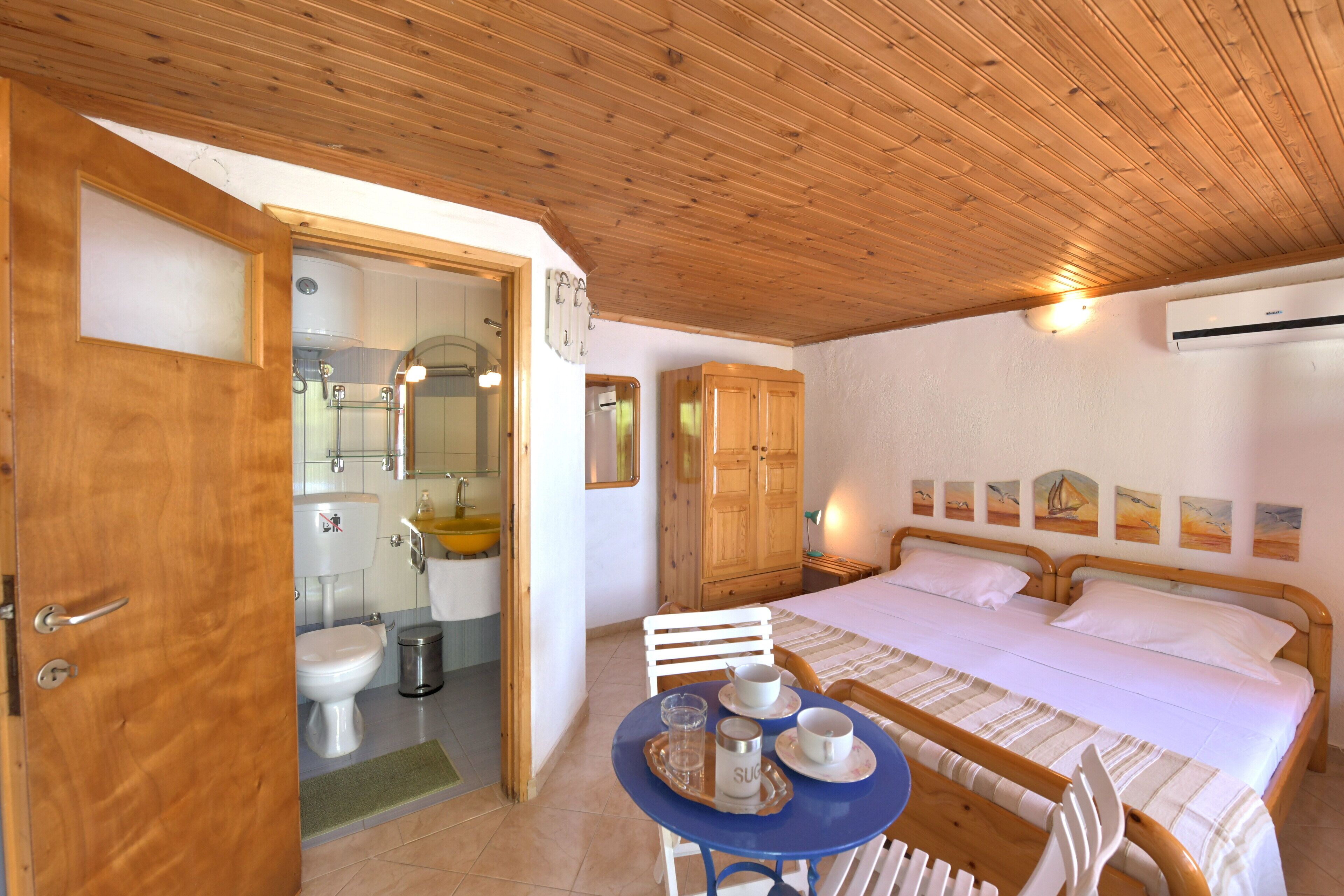 Une chambre avec un plafond en bois, un lit recouvert de draps blancs, une armoire en bois, une petite table avec un plateau, et une salle de bain avec des toilettes et un lavabo.