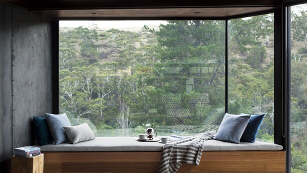 A modern room with a large window, a built-in bench with cushions, a small wooden stool, and a view of a forested area.