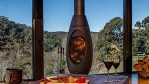 A rustic wooden table with a stoneware pot, a cutting board with cheese, grapes, and a red pepper, and two glasses of red wine.