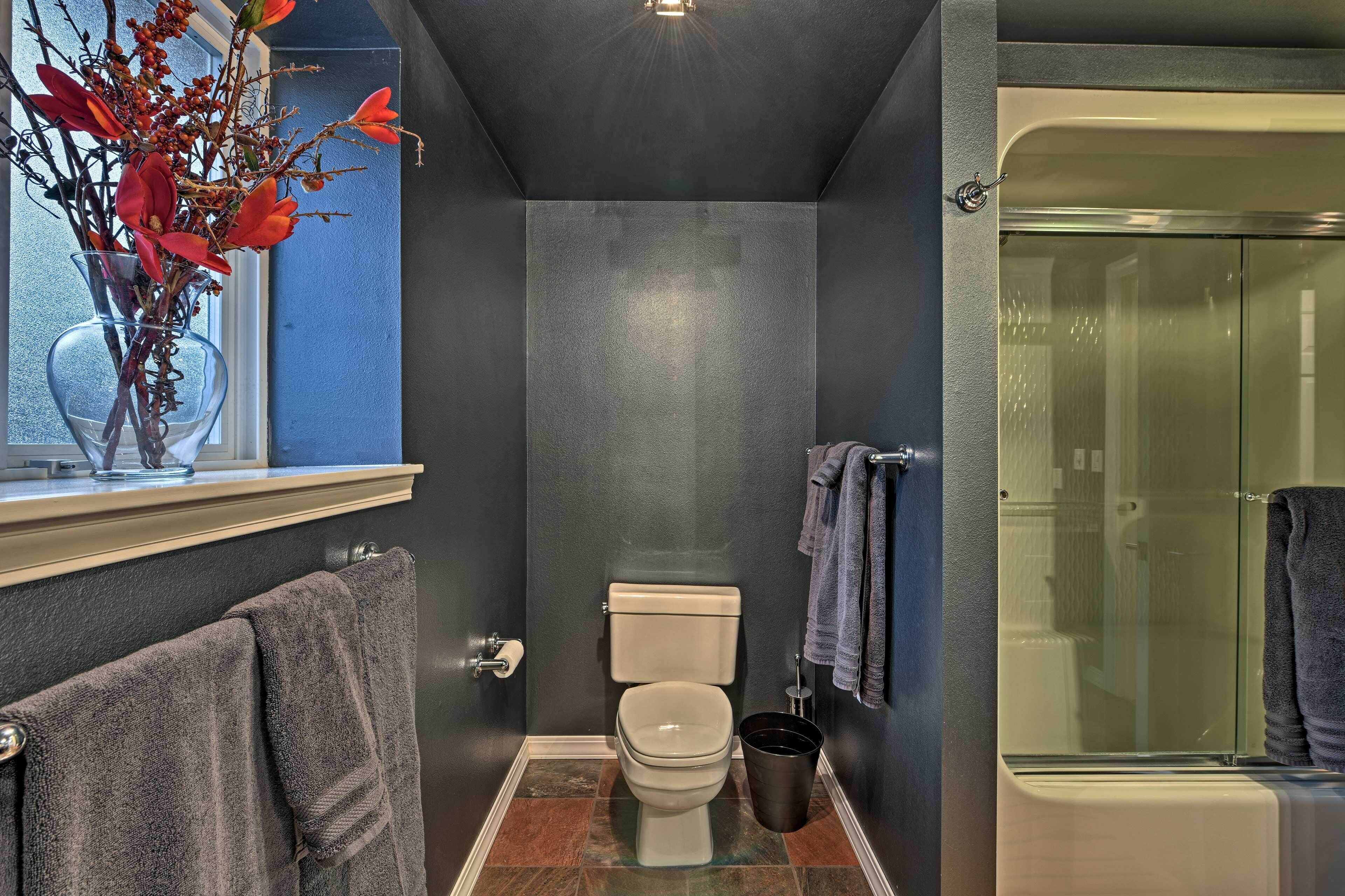 Un baño con inodoro, cabina de ducha de vidrio y un florero con flores rojas en el alféizar de una ventana.