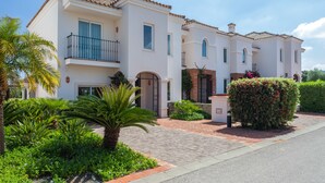 A two-story house with a tiled roof, a balcony, and a well-maintained garden with palm trees and shrubs. - Luxury 2 bedroom Frontline Golf Townhouse with shared pool in a gated community (Arcos de la Frontera)