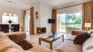 A spacious living room with a fireplace, a television, and a large window offering a view of the outdoors. - Luxury 2 bedroom Frontline Golf Townhouse with shared pool in a gated community (Arcos de la Frontera)