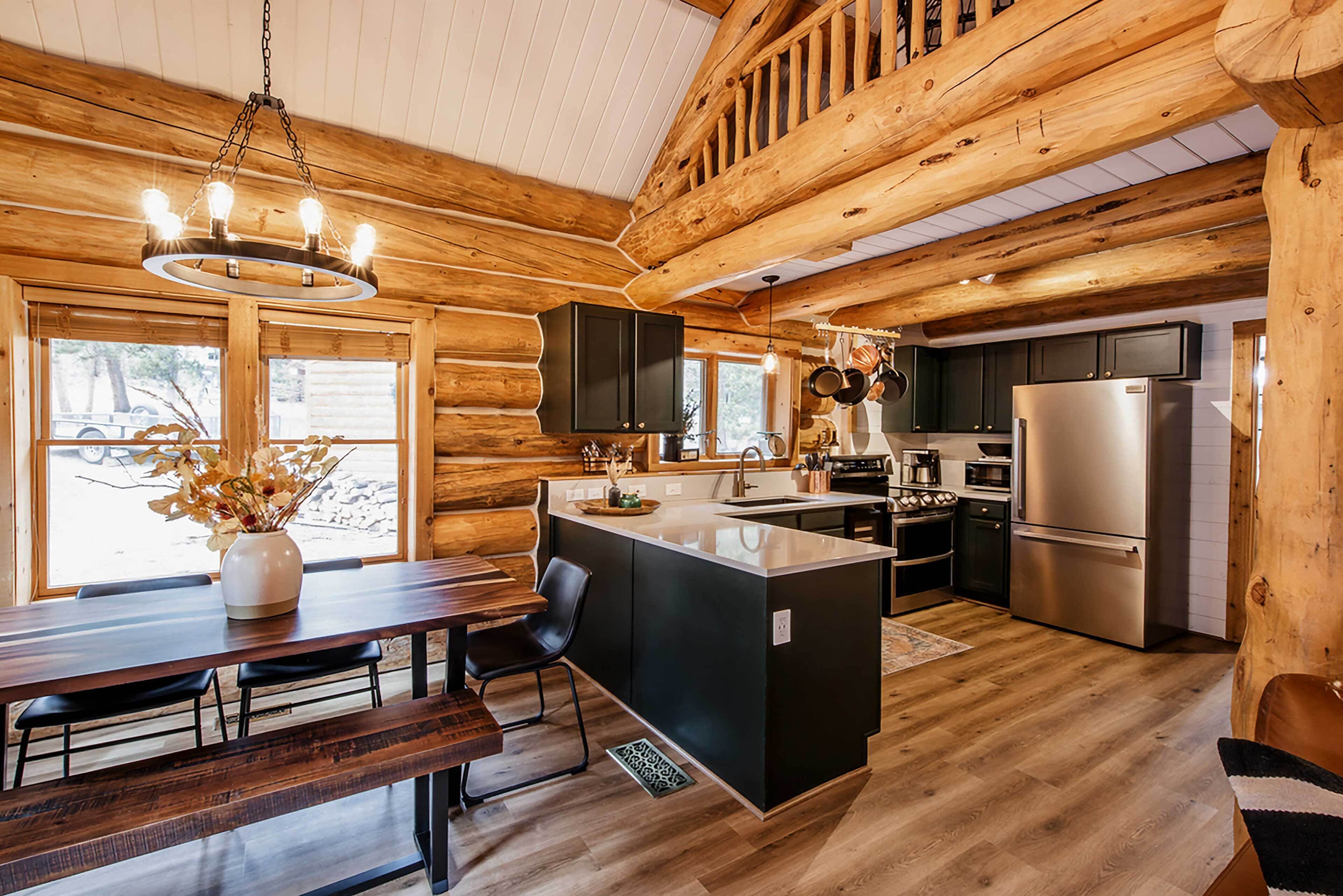 A log cabin interior with a wooden dining table, black chairs, a central island with a sink, and a stainless steel refrigerator.