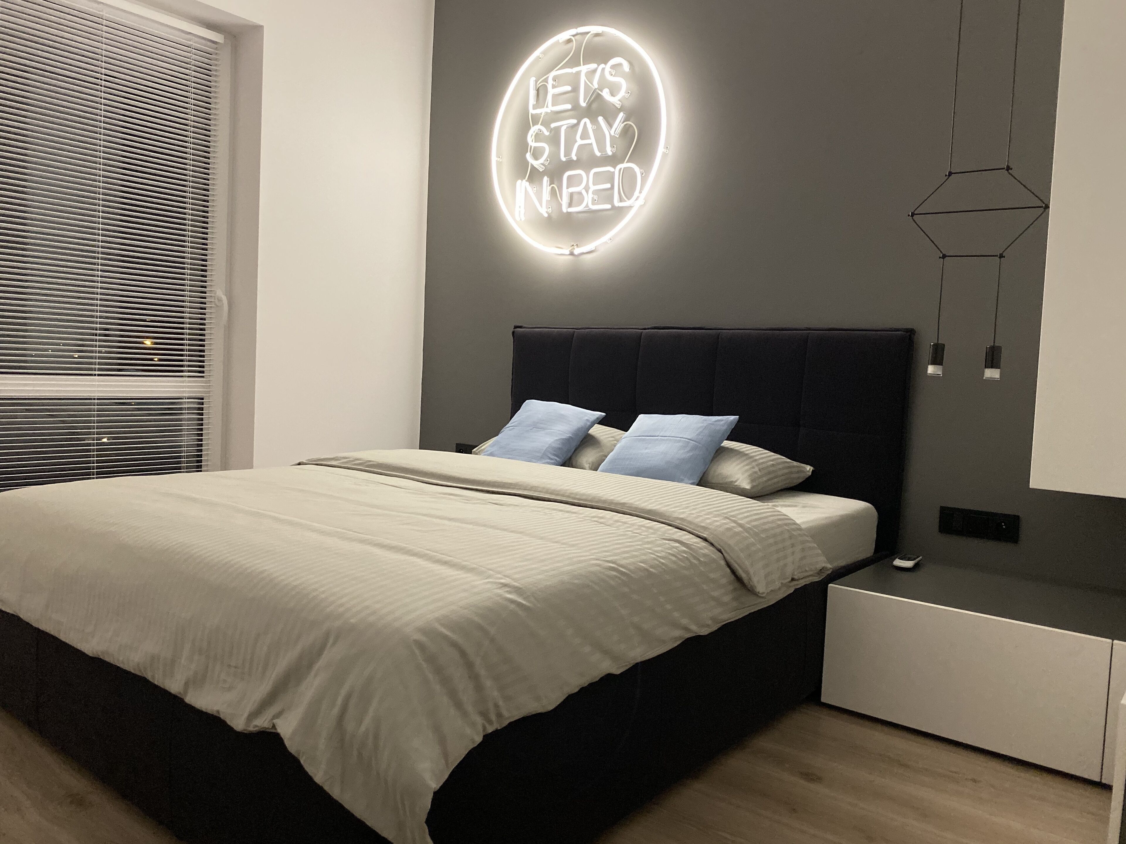 Un dormitorio moderno con cama, cabecero, mesita de noche, luz de pared y ventana con persianas.