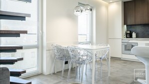 A modern kitchen with a white table and clear chairs, a built-in oven, and a window with blinds. - Premium Apartments in Wadowice (Wadowice)