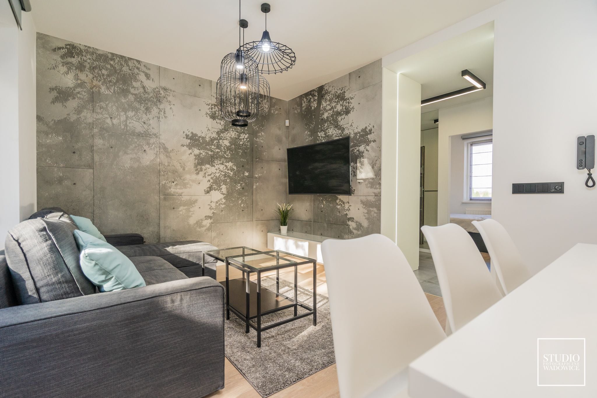 Una sala de estar moderna con un sofá gris, una mesa de centro de vidrio y un televisor montado en una pared con textura.