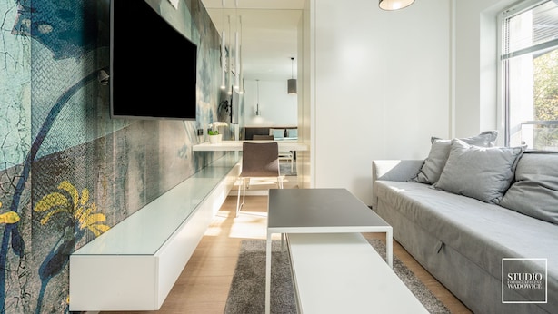 A modern living room with a flat-screen TV mounted on a textured wall, a grey sofa, a white coffee table, and a dining area in the background. - Premium Apartments in Wadowice (Wadowice)