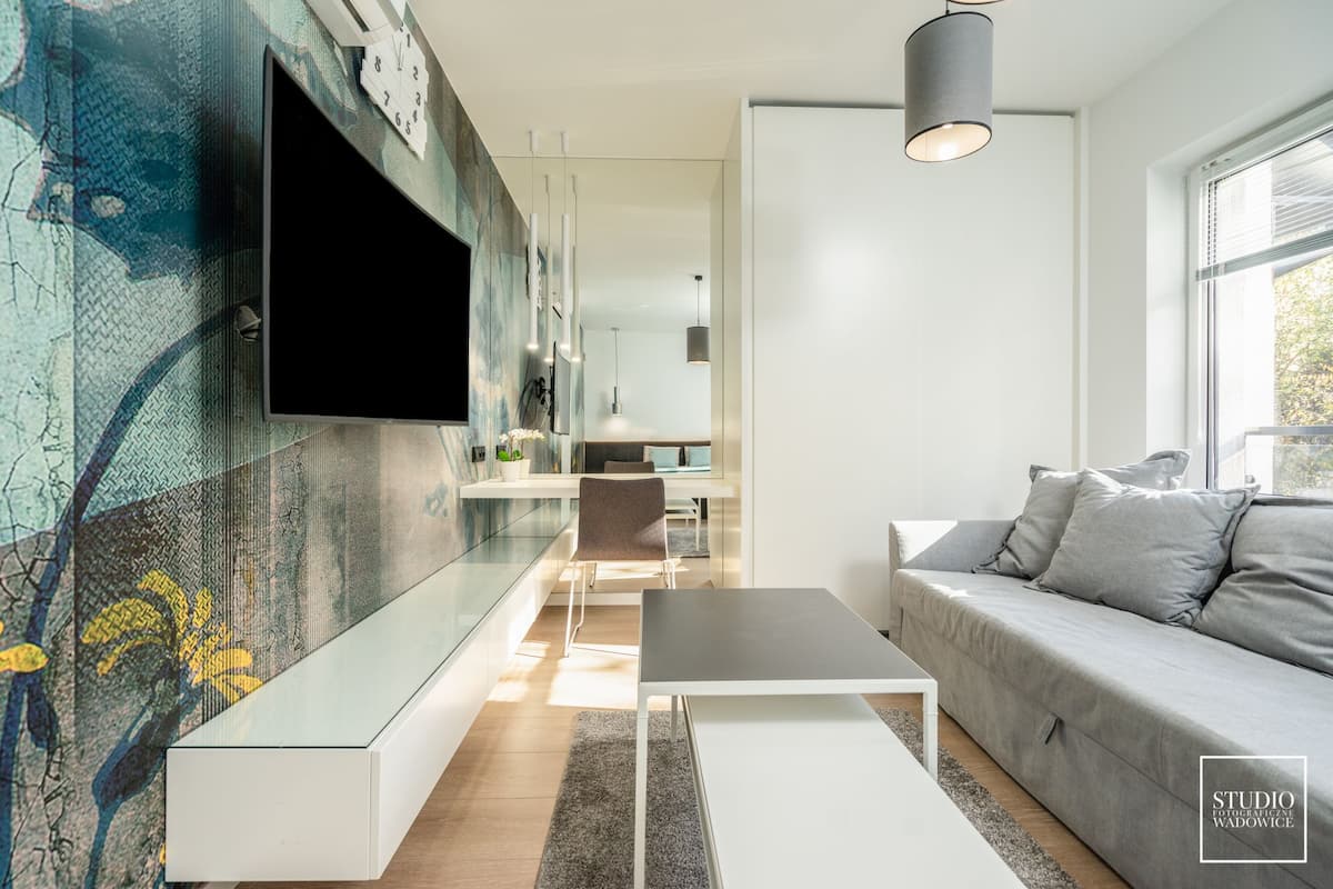 A modern living room with a flat-screen TV mounted on a textured wall, a grey sofa, a white coffee table, and a dining area in the background.