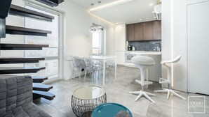 A modern kitchen with white cabinets, a dining area with a glass table and chairs, and a staircase with dark wood steps. - Premium Apartments in Wadowice (Wadowice)