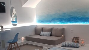 A modern bedroom with a large bed, a seating area with a sofa and pillows, a desk with a chair, and a wall-mounted mirror. - Amera Suites (Santorini)
