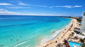 A beach with clear turquoise waters, a sandy shore, and a resort area with buildings and a swimming pool.