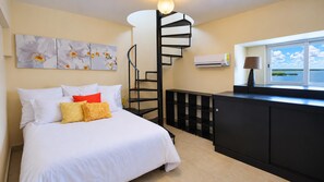 A bedroom with a bed, a black spiral staircase, a desk with a lamp, and a window with a view of the sky and water.