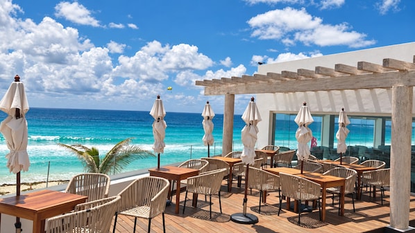An outdoor seating area with tables and chairs, overlooking a beach with the ocean and a clear sky.