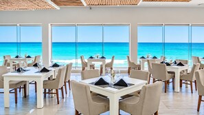 A restaurant with tables and chairs arranged for dining, large windows offering a view of the ocean.