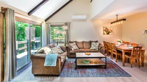 A spacious living room with a large window, a cozy sofa, a dining table with chairs, and a ceiling with exposed beams.