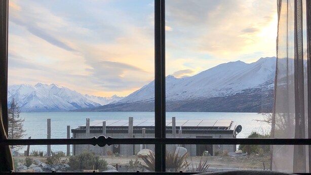 A view through a window of a mountainous landscape with a lake, a wooden fence, and a building in the foreground.