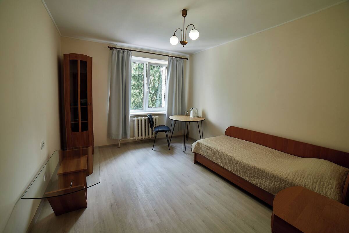 A bedroom with a bed, a glass table, a chair, a radiator, and a window with curtains.