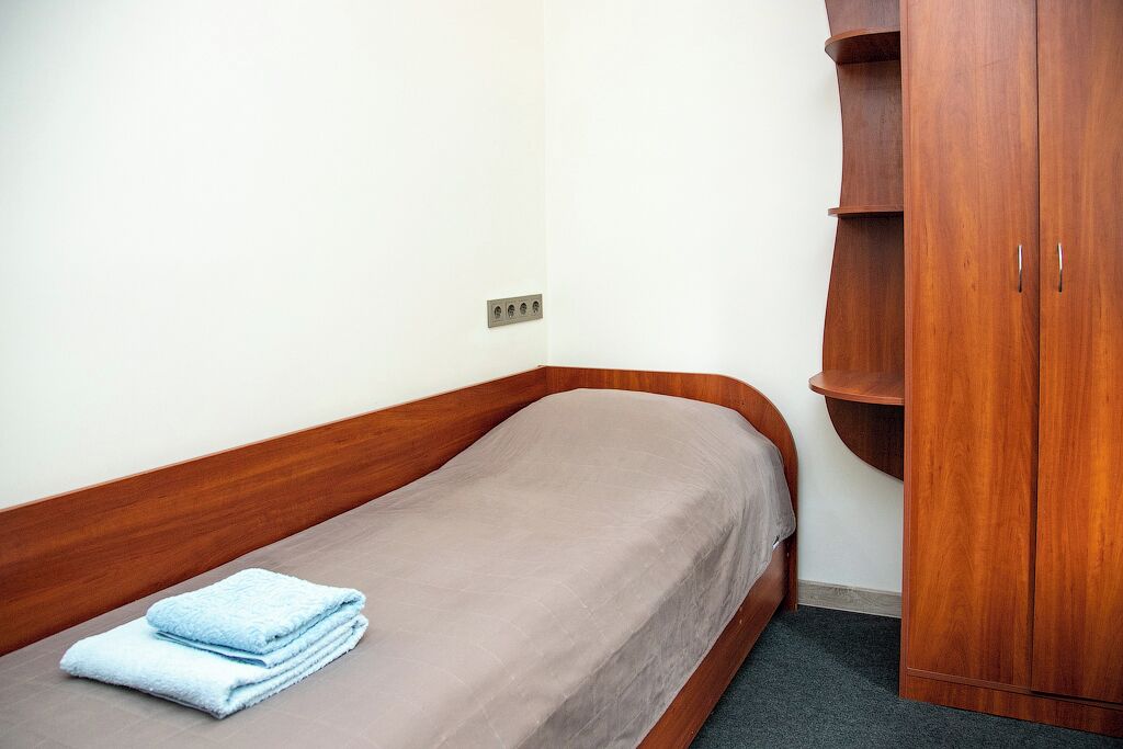 A single bed with a wooden headboard and a brown mattress, a blue towel on the bed, and a wooden wardrobe with open doors.