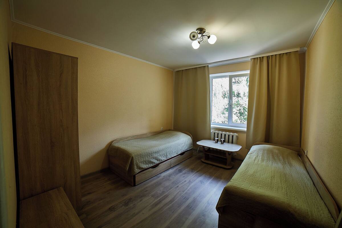 A small, single-room accommodation with two single beds, a small table, and a window with curtains.